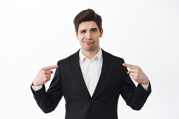 L'uomo insicuro e scettico in abito formale nero indica se stesso aggrotta le sopracciglia e sembra dubbioso su sfondo bianco