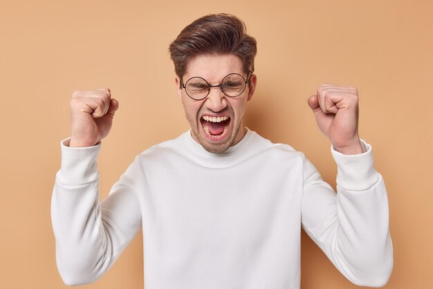 L'uomo irritato e indignato esprime emozioni negative tiene i pugni chiusi urla forte tiene la bocca aperta vestito con un maglione casual isolato su sfondo beige essendo infuriato. vi odio tutti