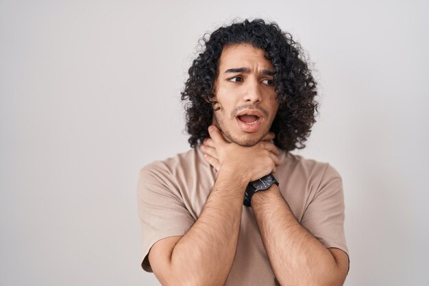 L'uomo ispanico con i capelli ricci in piedi su sfondo bianco grida soffocare perché strangolamento doloroso. problema di salute. concetto di asfissia e suicidio.