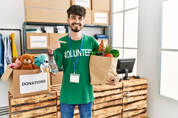 L'uomo ispanico con la barba che indossa una maglietta volontaria alle donazioni punta il dito verso un sé sorridente felice e orgoglioso