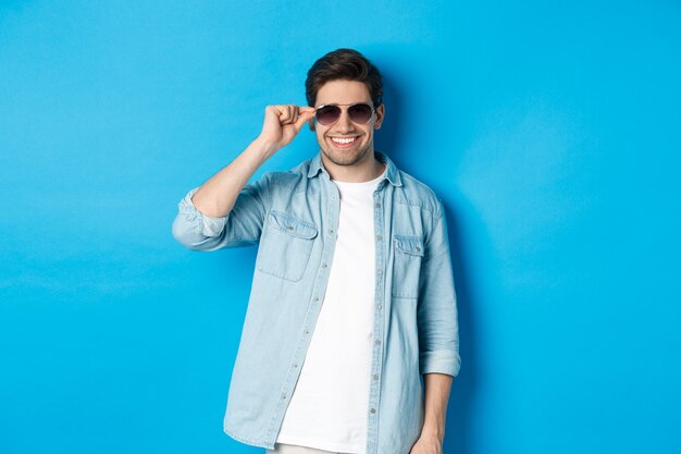 L'uomo macho fiducioso indossa gli occhiali da sole, sembra fresco e sfacciato, in piedi su sfondo blu