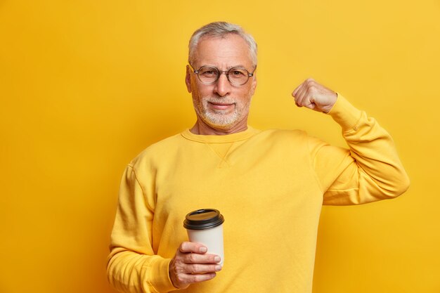 L'uomo maturo serio e sicuro di sé alza il braccio e mostra che il bicipite è forte e potente tiene la tazza di caffè di carta indossa un maglione casual isolato sopra il muro giallo