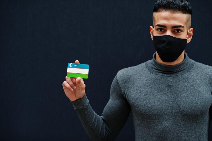 Foto gratuito l'uomo mediorientale con il collo alto grigio e la maschera nera per la protezione del viso mostra lo sfondo isolato della bandiera dell'uzbekistan