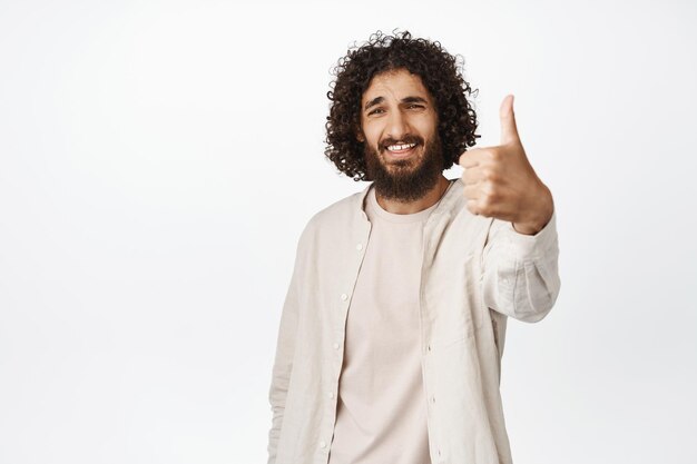 L'uomo mediorientale sorridente che mostra i pollici in su come e approva la lode smth buona raccomandando il fondo bianco del prodotto