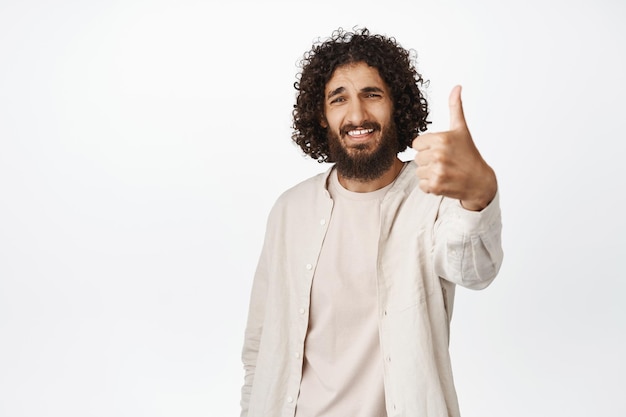 L'uomo mediorientale sorridente che mostra i pollici in su come e approva la lode smth buona raccomandando il fondo bianco del prodotto