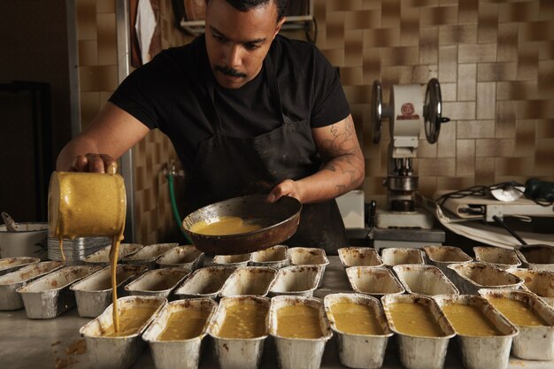 L'uomo nero versa una gustosa pastella per torta liquida dal misurino a speciali stampi realizzati con lamina metallica prima della cottura in forno. Processo di cottura professionale in panetteria artigianale