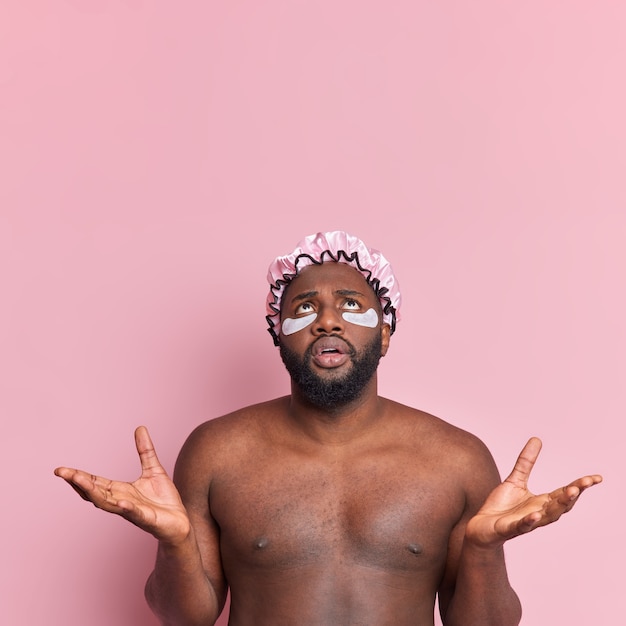 L'uomo perplesso allarga le palme con sconcerto concentrato sopra gli stand senza camicia indossa cappello da bagno e toppe di idrogel isolate sul muro rosa