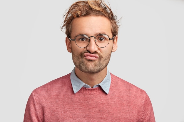 L'uomo perplesso e scontento curva le labbra, guarda dubbioso nella telecamera, prova esitazione, vestito con un maglione rosa, ha i capelli ricci, posa contro il muro bianco