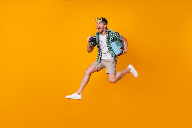 L'uomo positivo con la maschera subacquea in testa scatta foto con la fotocamera retrò. Ragazzo in pantaloncini e maglietta verde che salta con la valigia sullo spazio arancione.