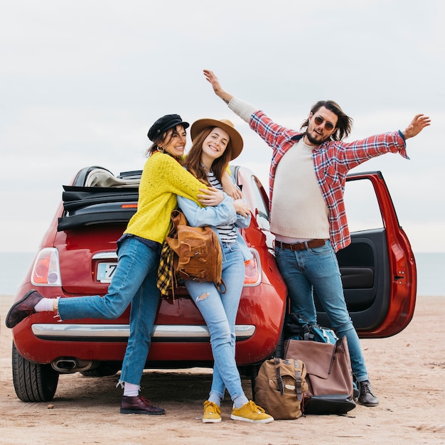 L&#39;uomo positivo con le mani alzate si avvicina ad abbracciare le donne e l&#39;automobile sulla spiaggia