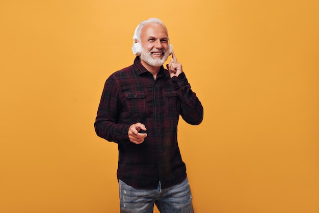 L'uomo positivo in camicia a quadri ascolta la musica in cuffia Adulto sorridente con barba grigia in posa su sfondo giallo isolato