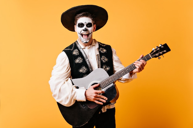 L'uomo positivo in sombrero canta una serenata. Ragazzo attivo con la chitarra nelle sue mani in posa sulla parete gialla.