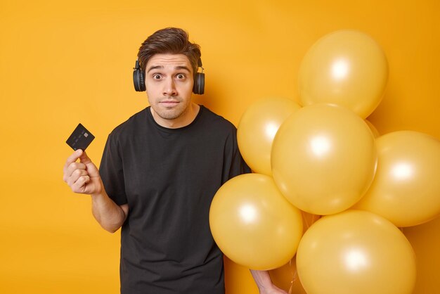 L'uomo sbalordito si prepara per la festa vestito con una maglietta nera casual tiene una carta bancaria e un mazzo di palloncini ascolta la traccia audio tramite le cuffie pone su sfondo giallo Tempo per la festa