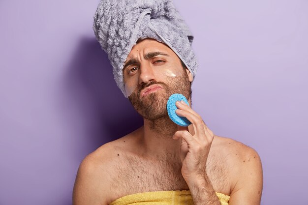 L'uomo serio rilassato con la barba incolta si asciuga la pelle del viso dopo la doccia, tiene la spugna cosmetica, avvolta in un asciugamano morbido