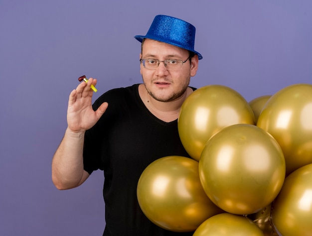 L'uomo slavo adulto compiaciuto con gli occhiali ottici indossa un cappello da festa blu con palloncini di elio che tengono un fischio di festa