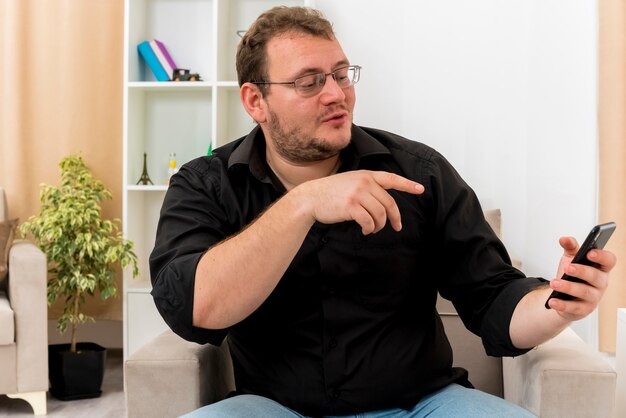 L'uomo slavo adulto compiaciuto con gli occhiali ottici si siede sulla poltrona guardando e indicando il telefono