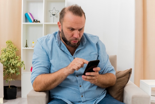 L'uomo slavo adulto scioccato si siede sulla poltrona indicando e guardando il telefono all'interno del soggiorno