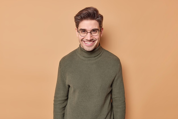 l'uomo sorride felice con i denti esprime emozioni allegre ascolta la storia divertente dell'interlocutore indossa occhiali rotondi e maglione isolato sul muro beige.