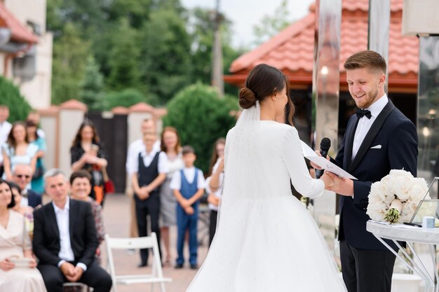 L'uomo sorridente dello sposo di vista laterale con il microfono che tiene la mano della sposa giura un giuramento sulla cerimonia di matrimonio all'aperto Ospiti felici sullo sfondo che si godono la bella coppia Moglie elegante in vestito gonfio