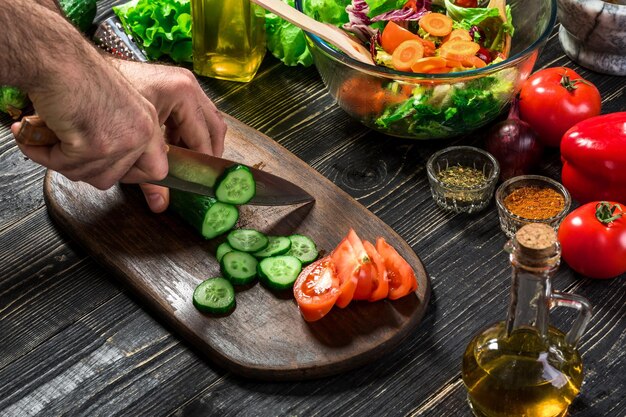 L'uomo sta tagliando il cetriolo su una tavola di legno nella cucina di casa sua per preparare l'insalata e mangiare la cena. Avvicinamento