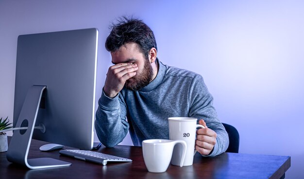 L'uomo stanco si siede davanti a un computer con una tazza di caffè con illuminazione colorata