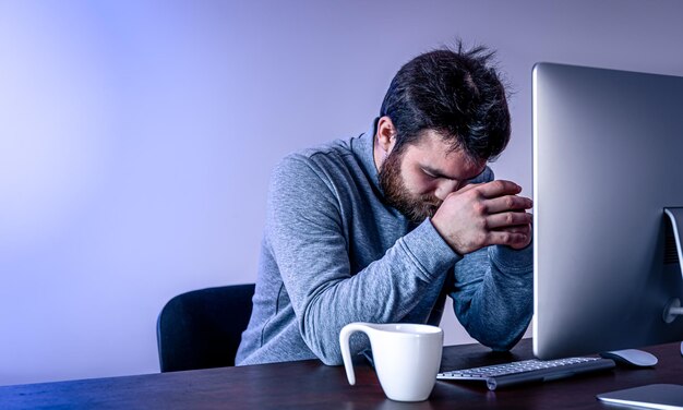 L'uomo stanco si siede davanti a un computer con una tazza di caffè con illuminazione colorata