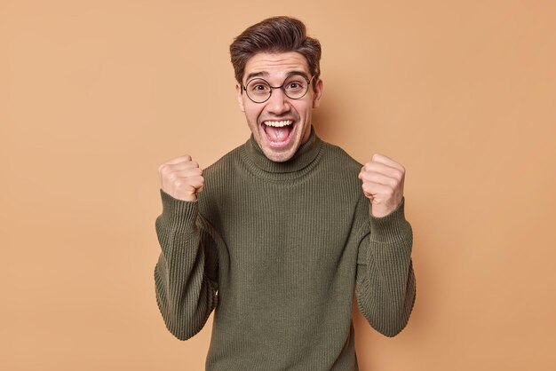 l'uomo stringe i pugni trionfa per un affare di successo o una grande notizia essendo gioioso ed entusiasta indossa maglione casual e occhiali pone contro il beige cattura il sogno