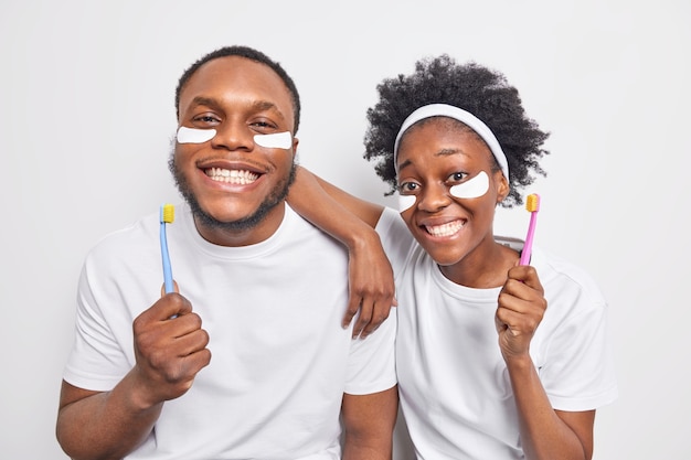 l'uomo tiene gli spazzolini da denti puliti i denti si sottopone a procedure di bellezza applica cerotti sotto gli occhi sentirsi felice gode della routine quotidiana mattutina isolata sul muro bianco.