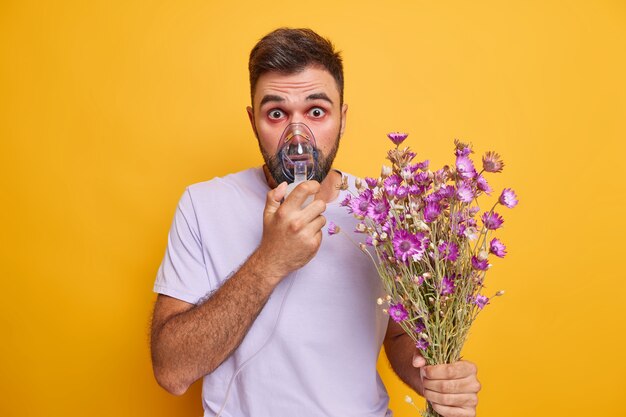 l'uomo usa l'inalatore dai fumi di polline dei fiori farmaci nei polmoni mehas occhi acquosi rossi tiene bouquet di fiori di campo vestiti casualmente isolati su giallo