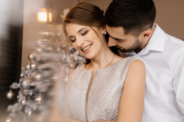 L&#39;uomo vestito elegante e la donna in abito d&#39;argento si abbracciano tenero in piedi davanti a un albero di Natale