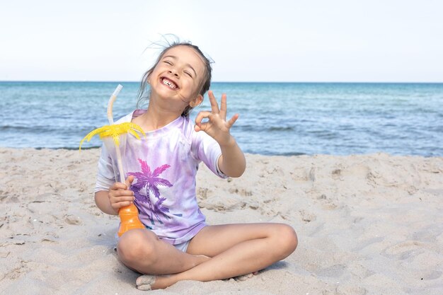 La bambina beve il succo seduto sulla sabbia vicino al mare