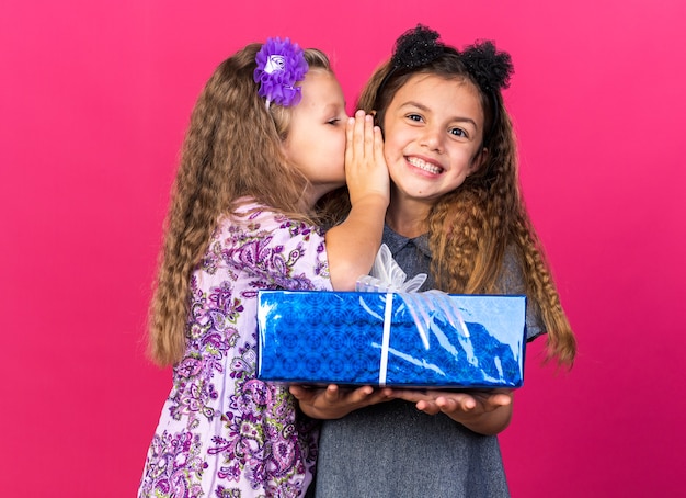 la bambina contenta sussurra all'orecchio della piccola ragazza caucasica sorridente che tiene il contenitore di regalo isolato sulla parete rosa con lo spazio della copia