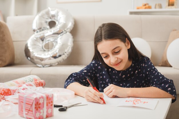 La bambina felice nel giorno della donna felice scrive su una cartolina seduta sul pavimento dietro il tavolino con regali in soggiorno