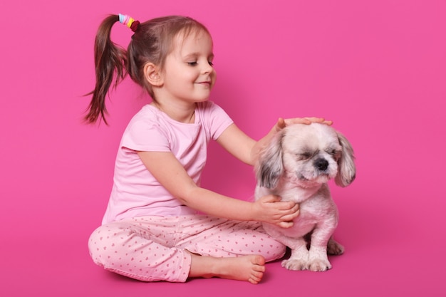 La bambina la nutre di pechinese mentre è seduta con le gambe incrociate sul pavimento. Adorabile bambino ama il suo animale domestico. Simpatico ragazzino sorridente guarda il suo cane, indossa camicia e pantaloni rosa, con code di cavallo. Concetto di bambini.