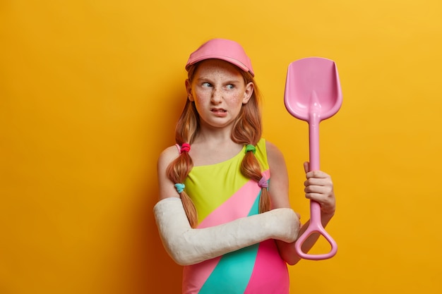 La bambina scontenta con i capelli rossi e le lentiggini guarda infelicemente la pala della sabbia, ha rovinato le vacanze estive a causa di un trauma, posa con un braccio rotto, necessita di un lungo trattamento, indossa un calco in gesso