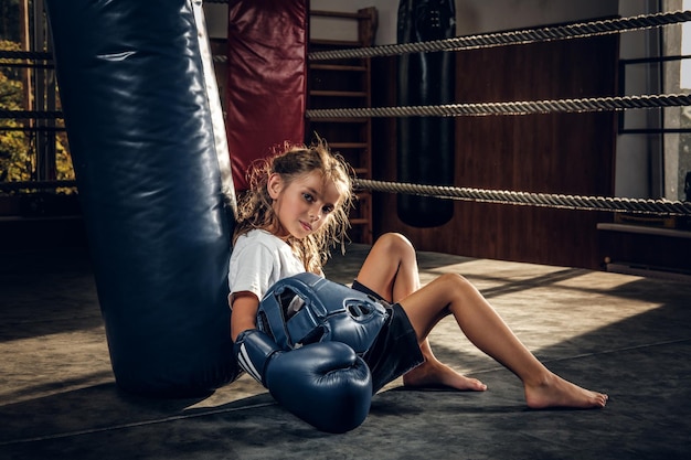 La bambina stanca con il casco blu sta riposando sull'anello accanto al sacco da boxe.