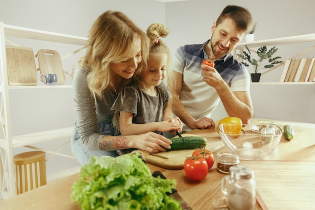 La bambina sveglia ed i suoi bei genitori stanno tagliando le verdure e sorridendo