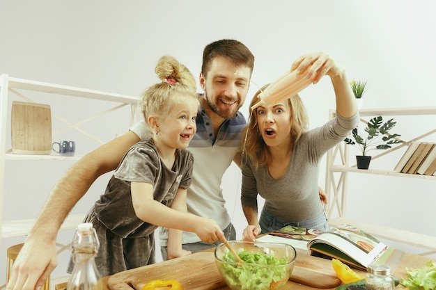 La bambina sveglia ei suoi bellissimi genitori stanno tagliando le verdure e sorridendo mentre fanno l'insalata in cucina a casa. Concetto di stile di vita familiare