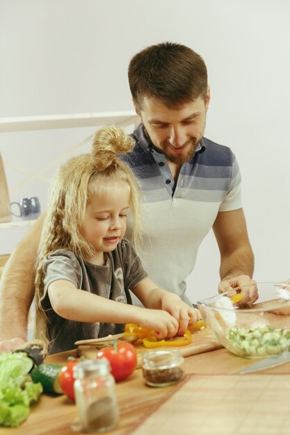 La bambina sveglia ei suoi bellissimi genitori stanno tagliando le verdure e sorridendo mentre fanno l'insalata in cucina a casa