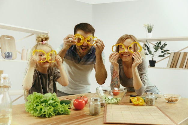 La bambina sveglia ei suoi bellissimi genitori stanno tagliando le verdure e sorridendo mentre fanno l'insalata in cucina a casa