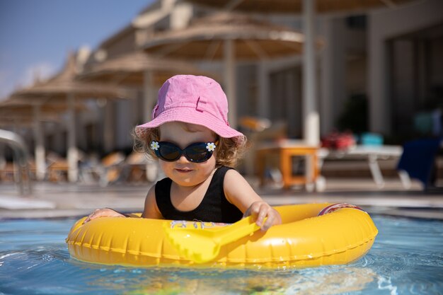 La bambina sveglia in un cappello e occhiali da sole gioca in piscina mentre era seduto in un cerchio di nuoto