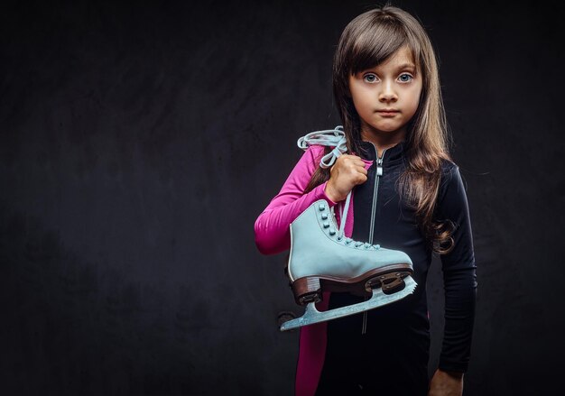 La bambina sveglia vestita di abbigliamento sportivo tiene i pattini da ghiaccio su una spalla. Isolato su uno sfondo scuro con texture.