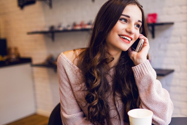 La battitura a macchina della donna scrive il messaggio sullo Smart Phone in un caffè moderno. Immagine potata di giovane ragazza graziosa che si siede ad una tavola con caffè o cappuccino facendo uso del telefono cellulare.