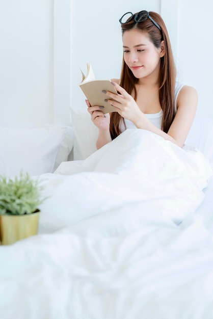La bella donna asiatica attraente gode di lettura del libro sul letto Il ritratto della donna asiatica dei capelli lunghi gode della camera da letto bianca di attività del fine settimana