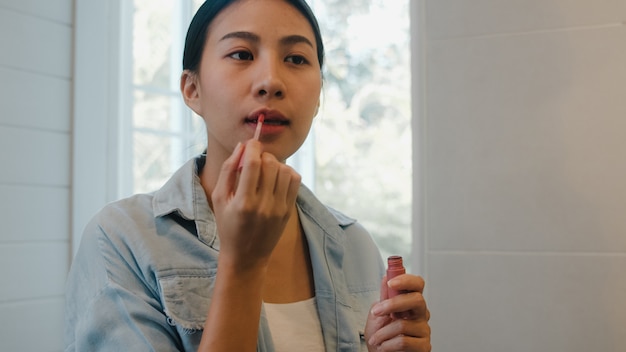 La bella donna asiatica che utilizza il rossetto compone nello specchio anteriore, femmina cinese felice che usando i cosmetici di bellezza per migliorarsi pronta a lavorare nel bagno a casa. Le donne dello stile di vita si rilassano a casa