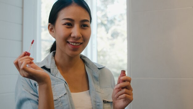 La bella donna asiatica che utilizza il rossetto compone nello specchio anteriore, femmina cinese felice che usando i cosmetici di bellezza per migliorarsi pronta a lavorare nel bagno a casa. Le donne dello stile di vita si rilassano a casa