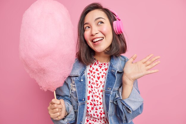 La bella donna asiatica con l'espressione sognante alza il palmo tiene il delizioso zucchero filato ascolta la traccia audio vestita con abiti alla moda isolati sul muro rosa. Concetto di divertimento per il tempo libero delle persone