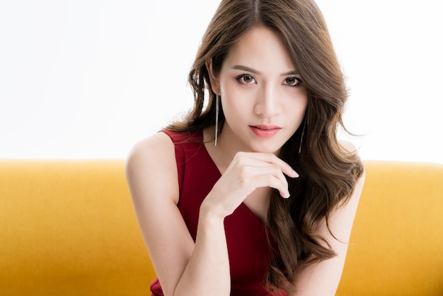 La bella donna asiatica di eleganza attraente in vestito rosso si siede e si rilassa la posa del modello sul servizio fotografico giallo dello studio del sofà