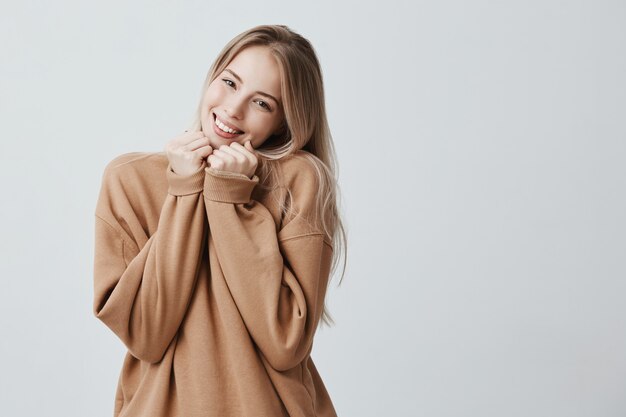 La bella donna bionda esprime emozioni felici, in maglione marrone, ha un ampio sorriso piacevole, felice di ricevere il presente. Il modello femminile estatico sorridente gode della vita, isolata