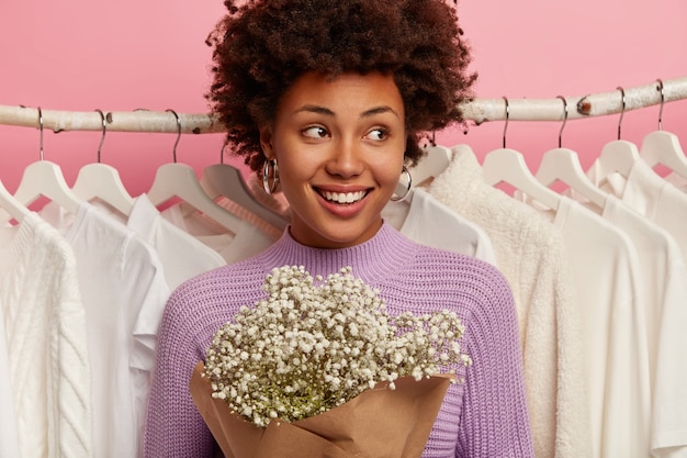 La bella donna compiaciuta ha i capelli ricci ha un sorriso a trentadue denti, indossa un maglione viola casual, tiene un bouquet, sta contro il maglione bianco sui rack